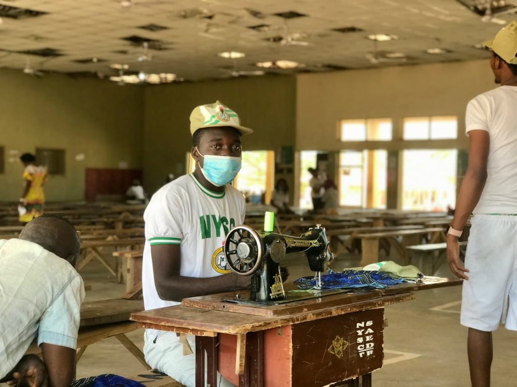 a-man-in-white-shirt-wearing-face-mask-while-sitting-near-the-sewing-machine-on-a-wooden-table-6802855 A young man sewing during the National Youth Service Corps program in Nigeria, promoting skill development.